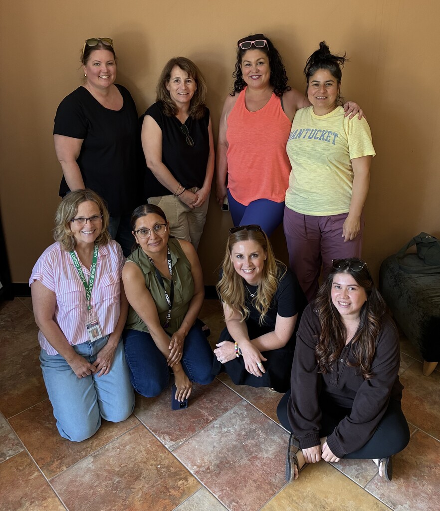 Nine women in casual clothing pose for a group photo. Half are seated and half are standing.