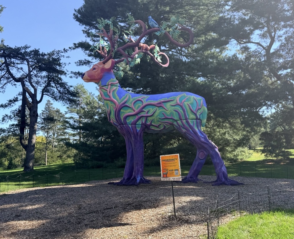 A large, vividly painted deer statue stands in a park with tall trees and grassy areas.