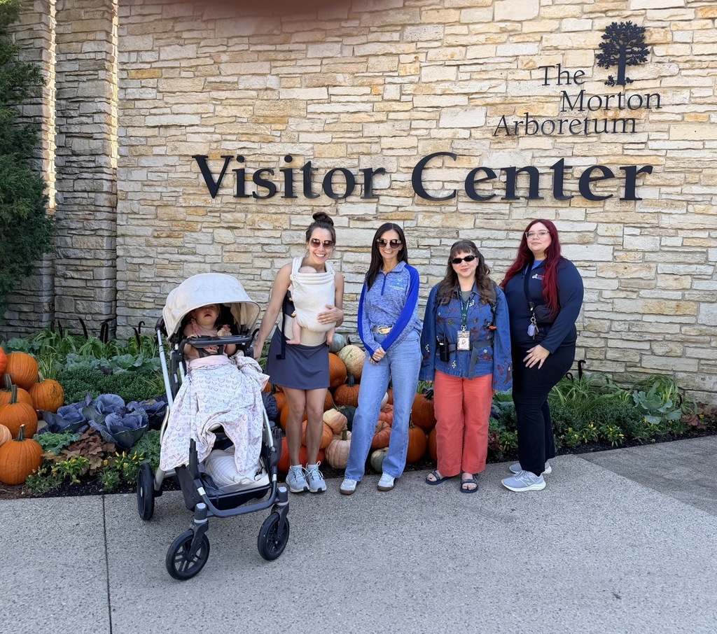 Five women, including one in a stroller, stand in front of a stone wall. The wall says "The Morton Arboretum Visitor Center" above them.