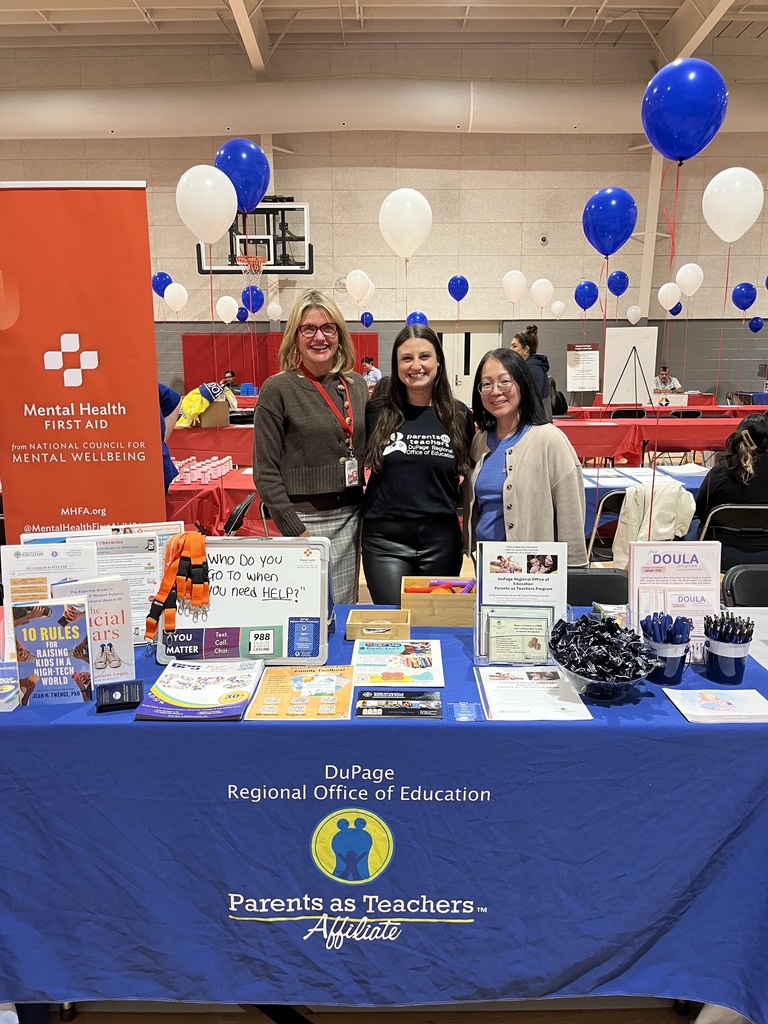 Three women stand behind a table with pamphlets, books, and balloons. A banner reads "Parents as Teachers."