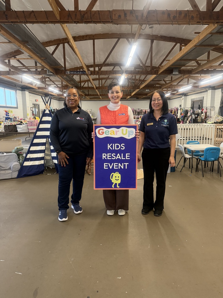 Three women stand near a sign in a room. The sign reads "Kids Resale Event." One woman holds the sign.