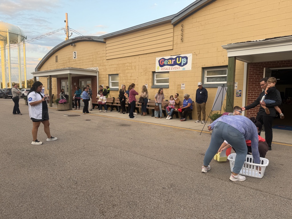 People gather outside a yellow building with a "Gear Up" sign. Some are standing, some sitting, and some carrying baskets.