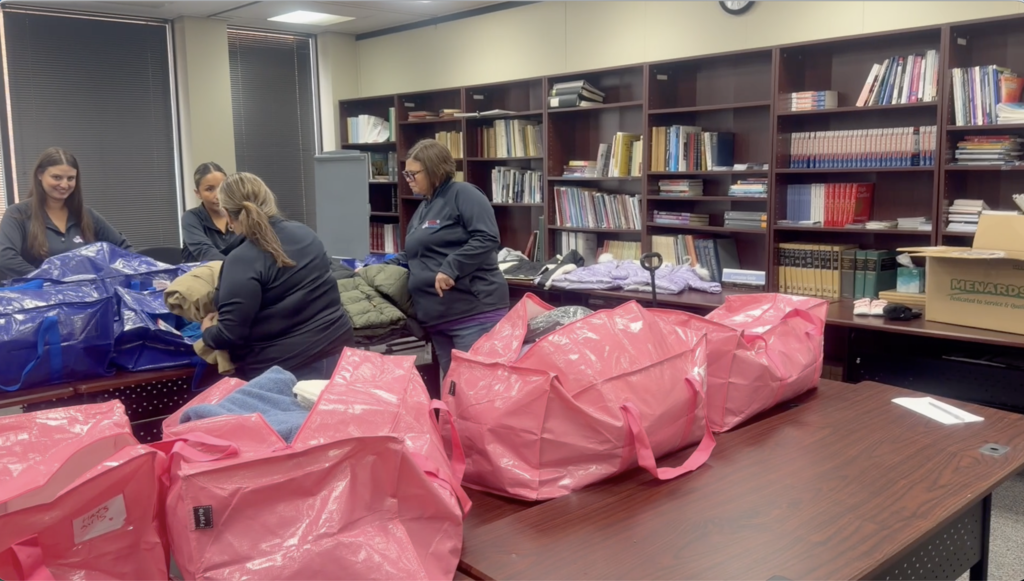 Five women in a library with shelves filled with books. They are sorting items into pink bags on a desk.