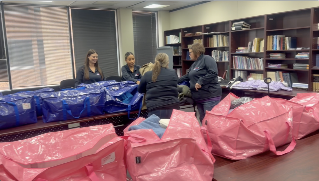 Four women prepare and pack bags in a room with bookshelves. Multiple bags in pink and blue.