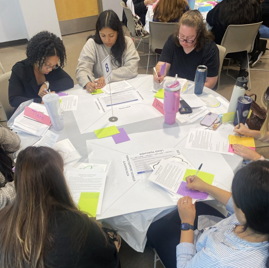 A group of women sitting around a table, writing on papers. Papers, pens, and water bottles are on the table.