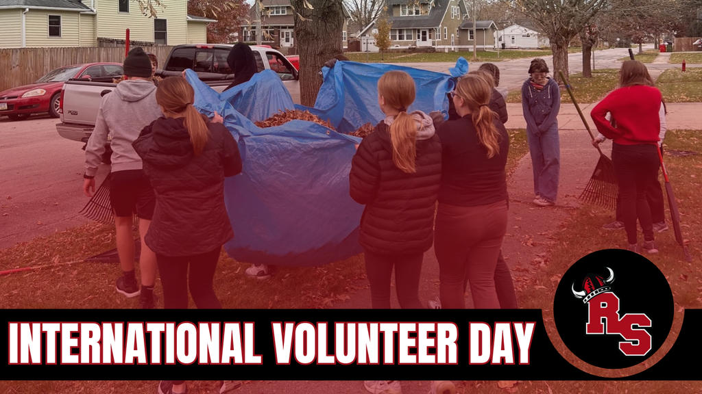Several middle school students wearing jackets work together outdoors to rake and collect fallen leaves into large blue tarps. They stand near a residential street lined with houses and trees that have lost most of their leaves. A red and black banner across the bottom reads ‘International Volunteer Day’ with the Roland-Story school logo on the right.