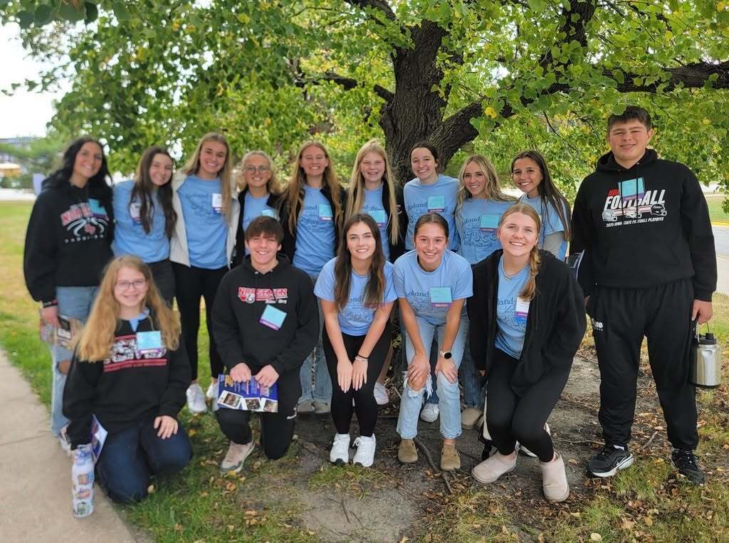 A group of people wearing matching blue shirts stands under a tree, with some sitting and others standing.