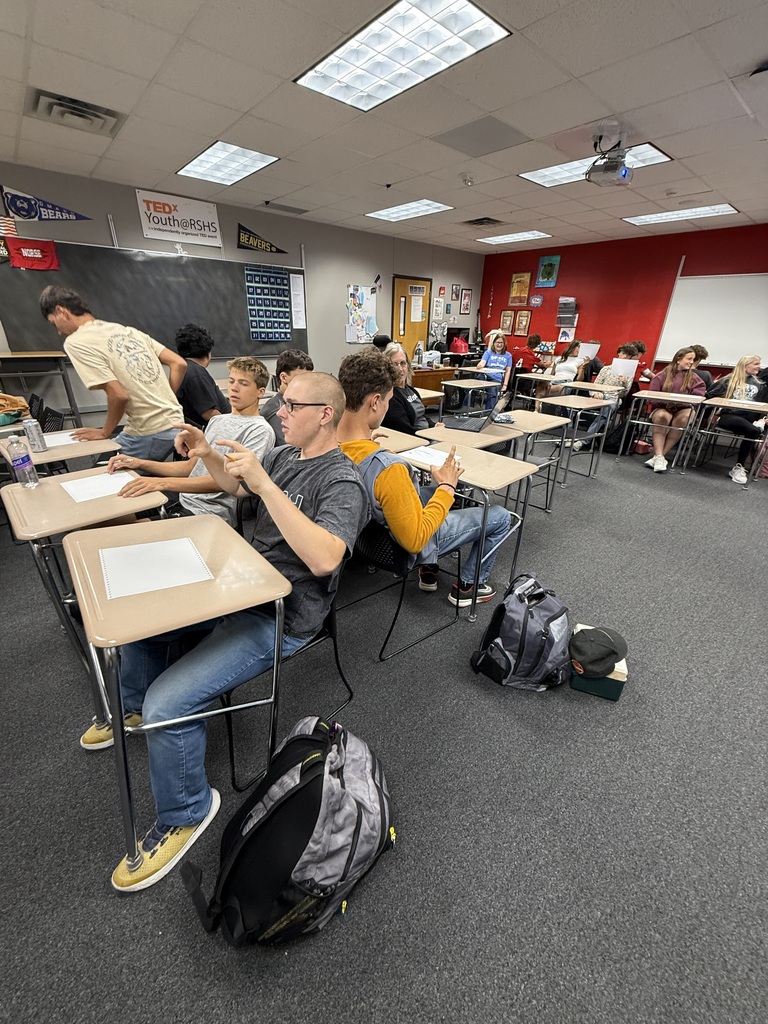 A classroom with desks and chairs. Students sit and stand. Backpacks are on the floor. Some wear glasses.