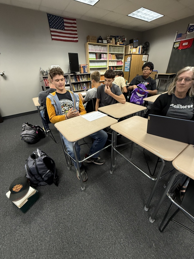 Several students seated at desks in a classroom. One student is working on a laptop.