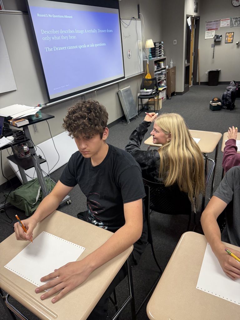 A classroom with a projector screen. A student writes on paper, and others sit at tables with papers and pencils.