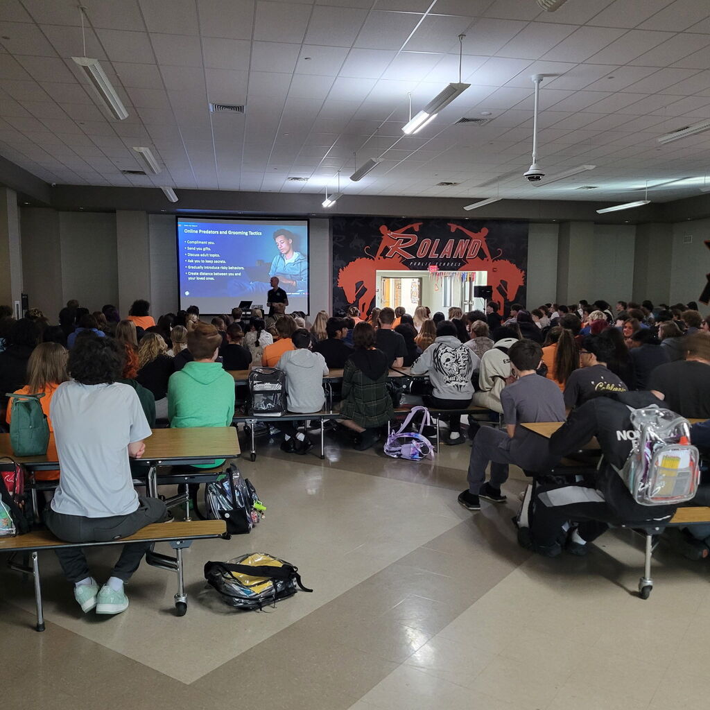 Two photos show Anthony Meter with Homeland Security speaking to Roland High School students about online safety, cell phone safety, social media awareness, and recognizing potential online dangers. Students are seated in the school cafeteria listening to the presentation.
