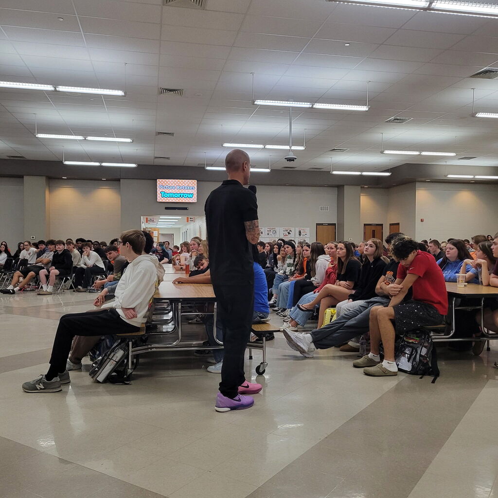 Two photos show Anthony Meter with Homeland Security speaking to Roland High School students about online safety, cell phone safety, social media awareness, and recognizing potential online dangers. Students are seated in the school cafeteria listening to the presentation.