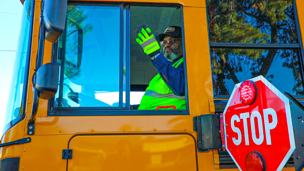 A bus driver waves out of the window while the stop sign is extended