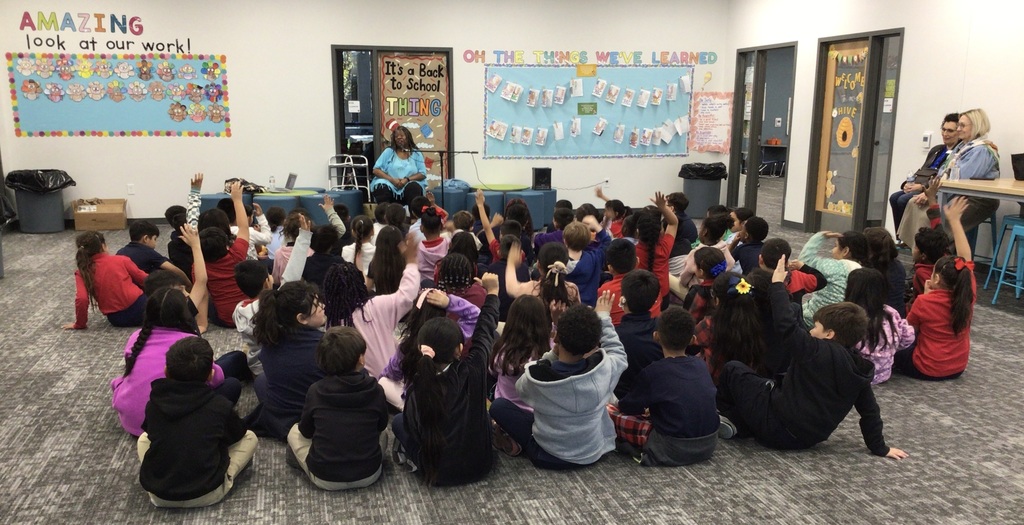 Students sitting on floor with hands raised listening to story telling
