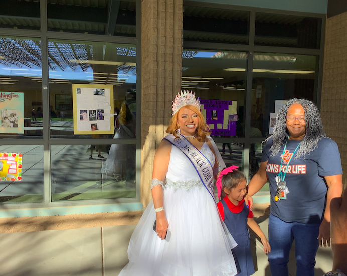 Lady in white gown and crown with sash, a female student with red bow in her hair, 1 teacher taking a picture together.