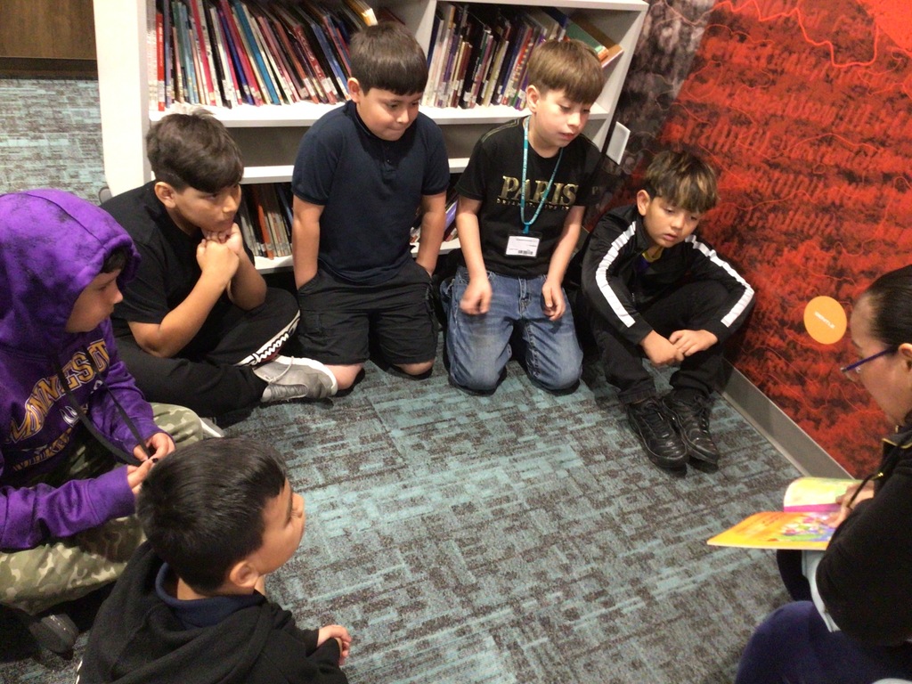 A lady in a black shirt is reading to 6 students sitting on the floor.