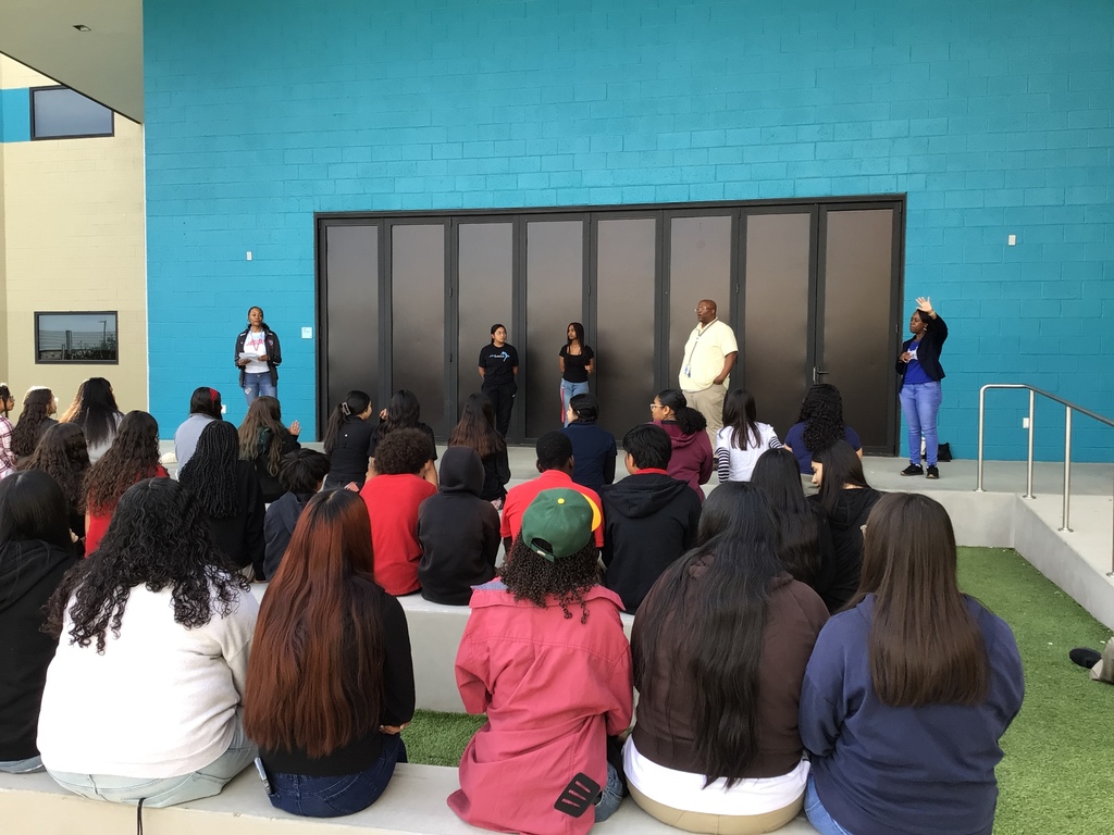 3 adults and 2 high school students on outside stage with a blue wall backdrop speaking to 8th graders.