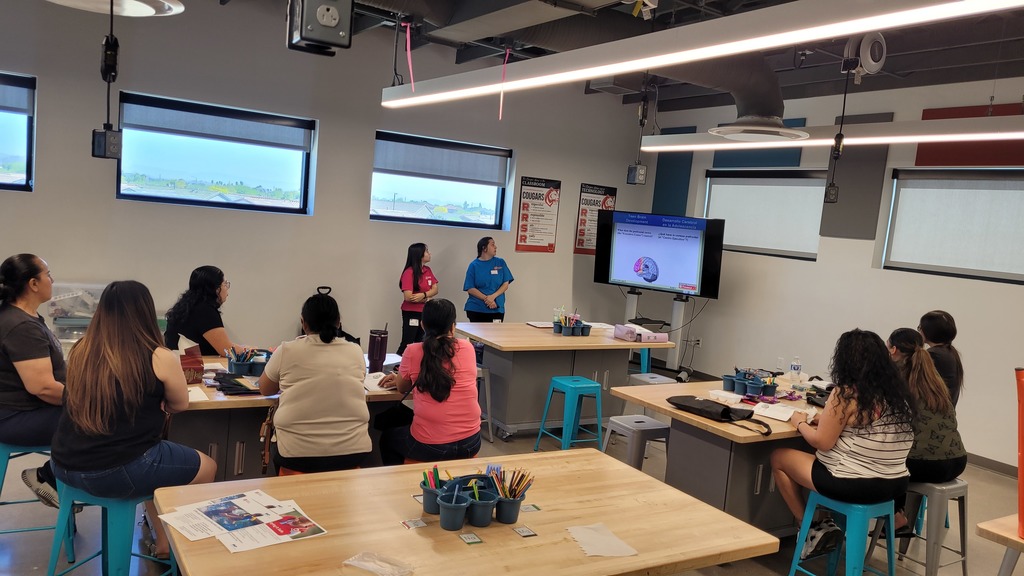 8 parents sitting on stools listening to 2 teachers.