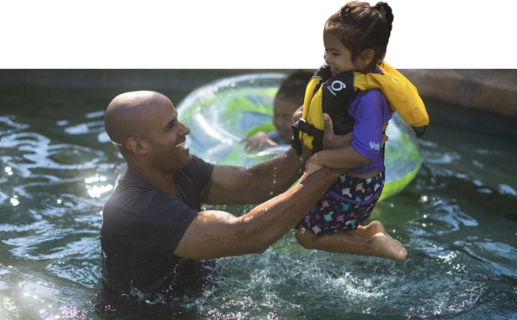 A man holding up a child wearing a life jacket while in the pool.
