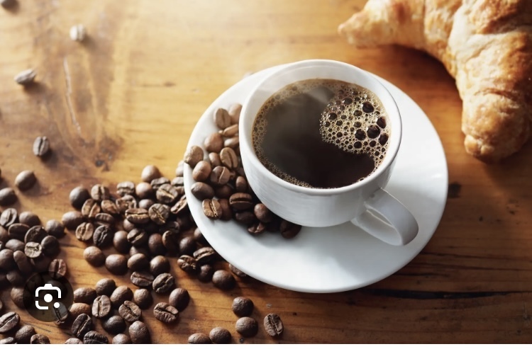 A white cup of black coffee on a saucer, surrounded by scattered coffee beans on a wooden table, with a croissant in the background.