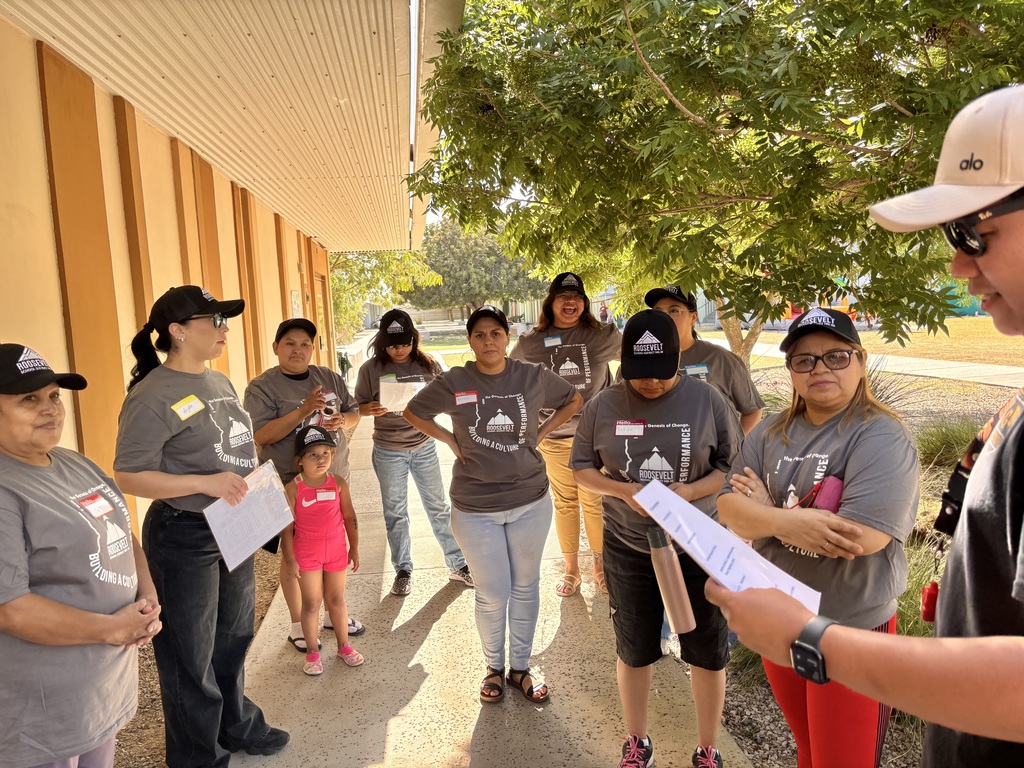 A group of parent volunteers wearing matching gray Roosevelt T-shirts and black Roosevelt caps gathers outside a school building during an orientation. A staff member in the foreground reviews information from a printed sheet while the volunteers listen. One young child in a bright pink outfit stands with the group. Trees provide shade along the walkway, creating a welcoming outdoor meeting space.
