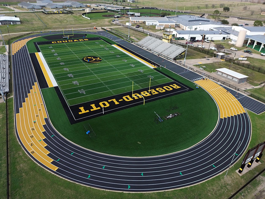 Aerial view of a high school football field with a black and green design, surrounded by a black running track with yellow accents. Bleachers are on one side.