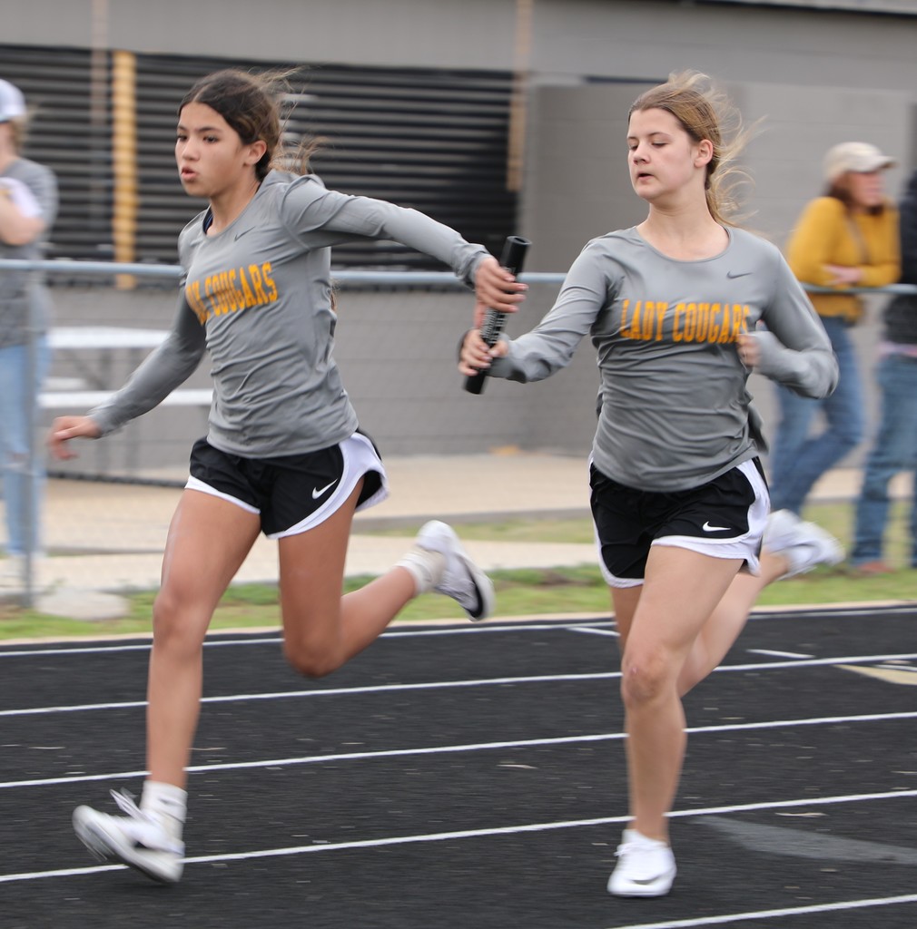 Two young female athletes in gray "Lady Cougars" shirts are running on a track, passing a baton in a relay race, conveying focus and teamwork.