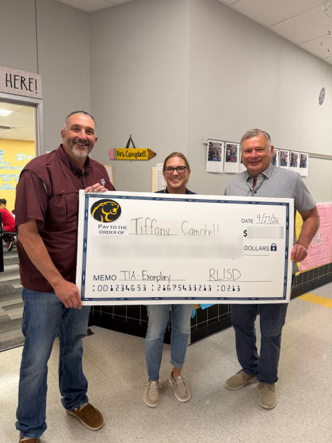 Three people stand in a school hallway holding a large, novelty check. The mood is celebratory and positive, with colorful decorations and student artwork on the walls.