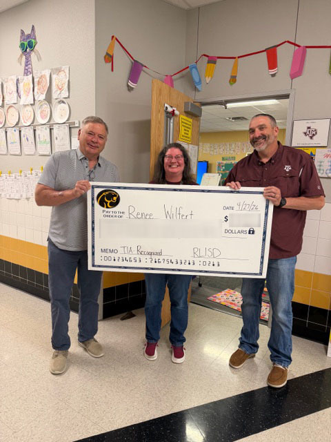Three people stand in a school hallway holding a large, novelty check. The mood is celebratory and positive, with colorful decorations and student artwork on the walls.