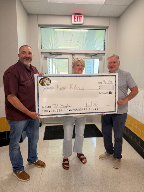 Three people stand in a school hallway holding a large, novelty check. The mood is celebratory and positive, with colorful decorations and student artwork on the walls.