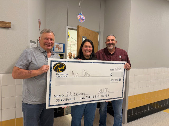 Three people stand in a school hallway holding a large, novelty check. The mood is celebratory and positive, with colorful decorations and student artwork on the walls.