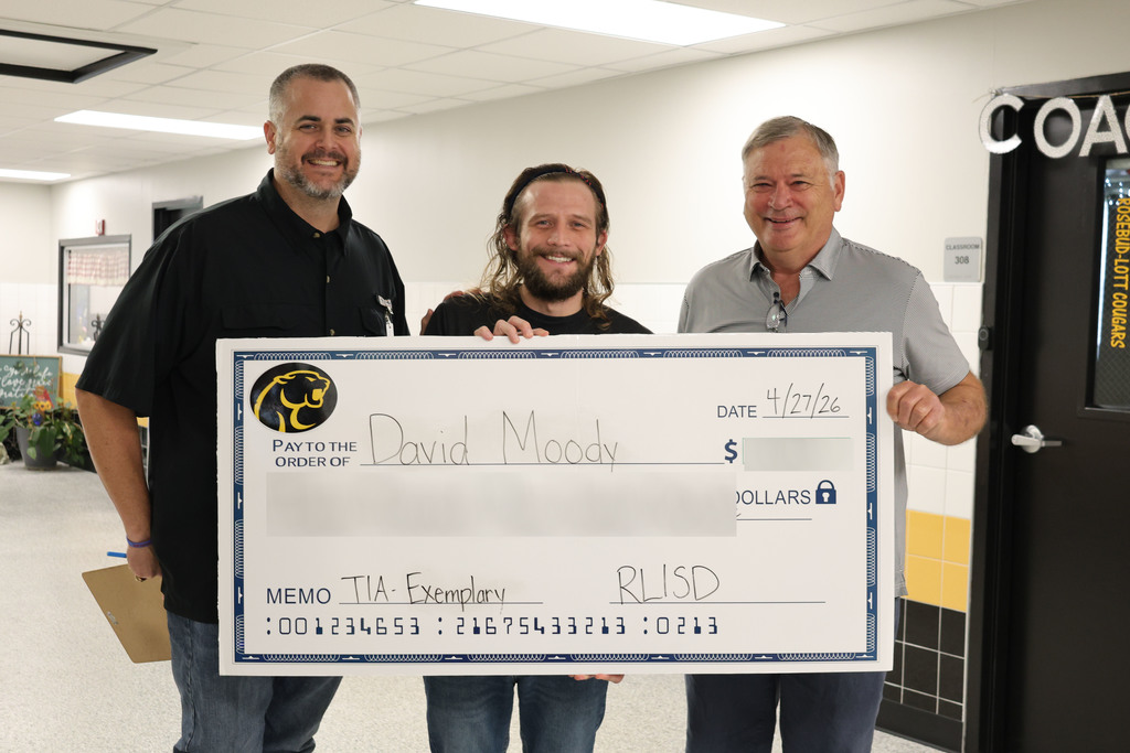Three people stand in a school hallway holding a large, novelty check. The mood is celebratory and positive, with colorful decorations and student artwork on the walls.