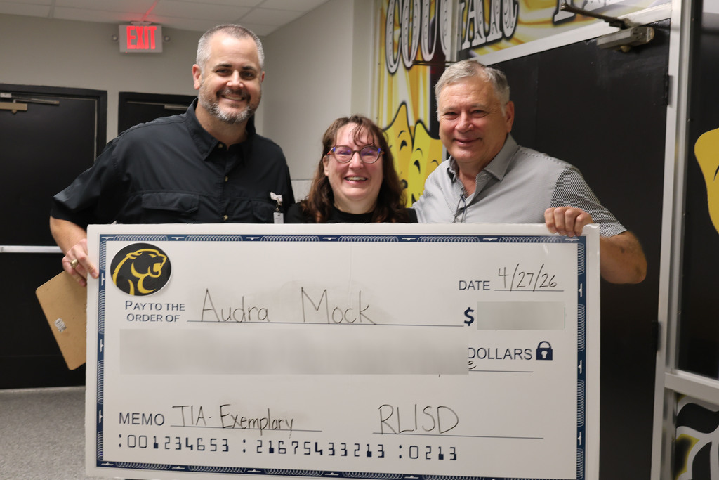 Three people stand in a school hallway holding a large, novelty check. The mood is celebratory and positive, with colorful decorations and student artwork on the walls.