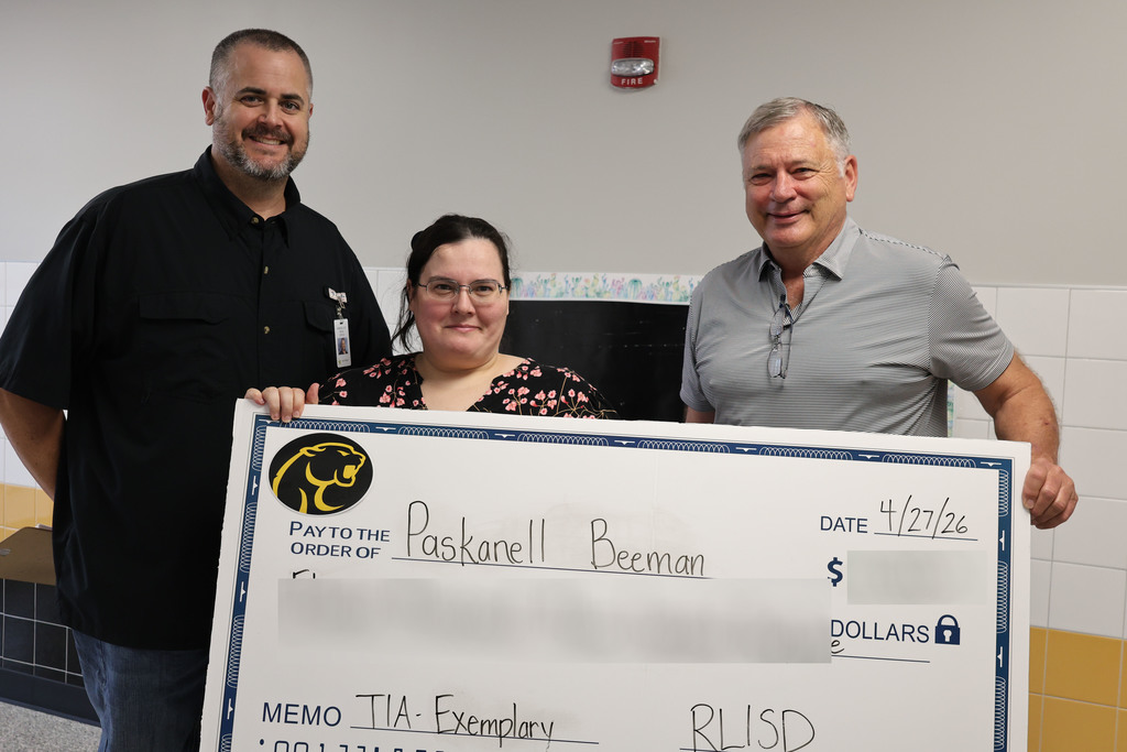 Three people stand in a school hallway holding a large, novelty check. The mood is celebratory and positive, with colorful decorations and student artwork on the walls.