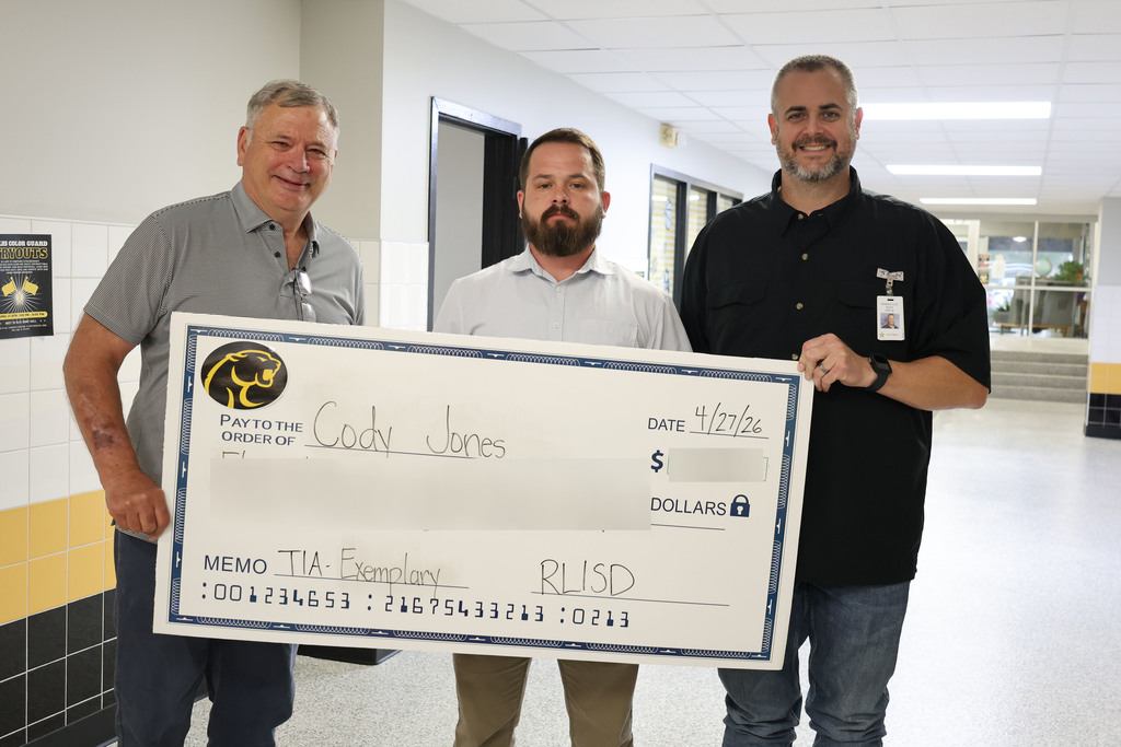 Three people stand in a school hallway holding a large, novelty check. The mood is celebratory and positive, with colorful decorations and student artwork on the walls.