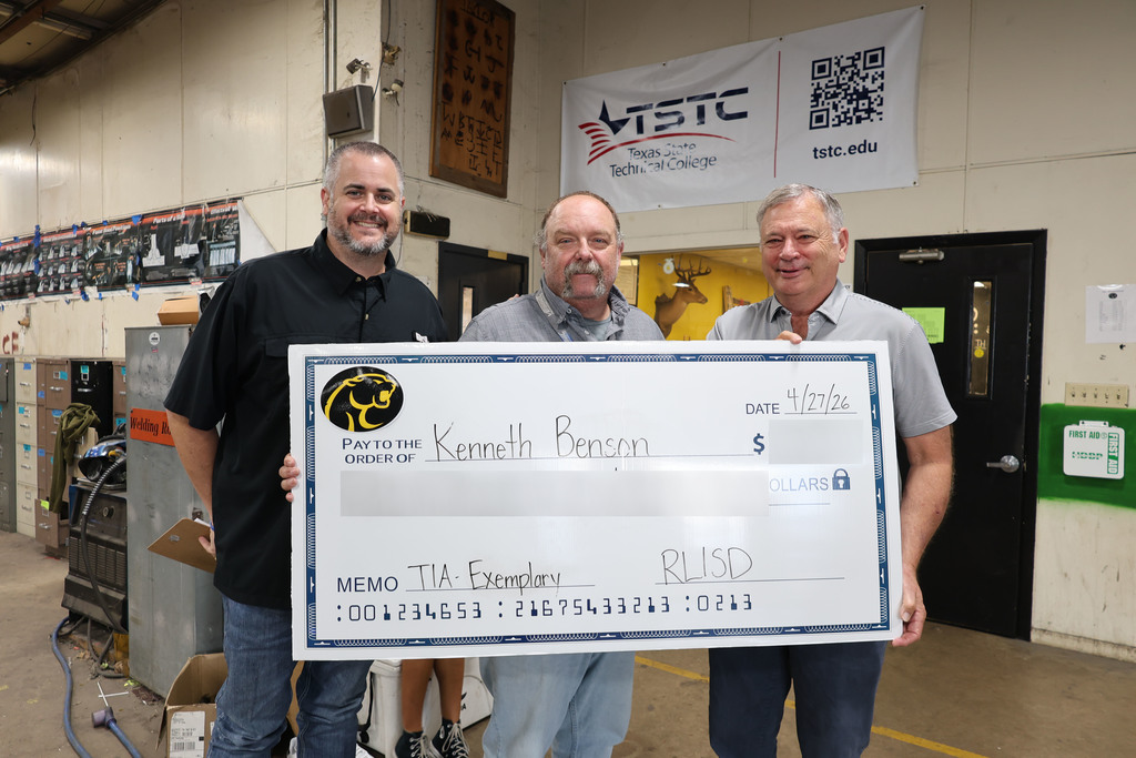 Three people stand in a school hallway holding a large, novelty check. The mood is celebratory and positive, with colorful decorations and student artwork on the walls.