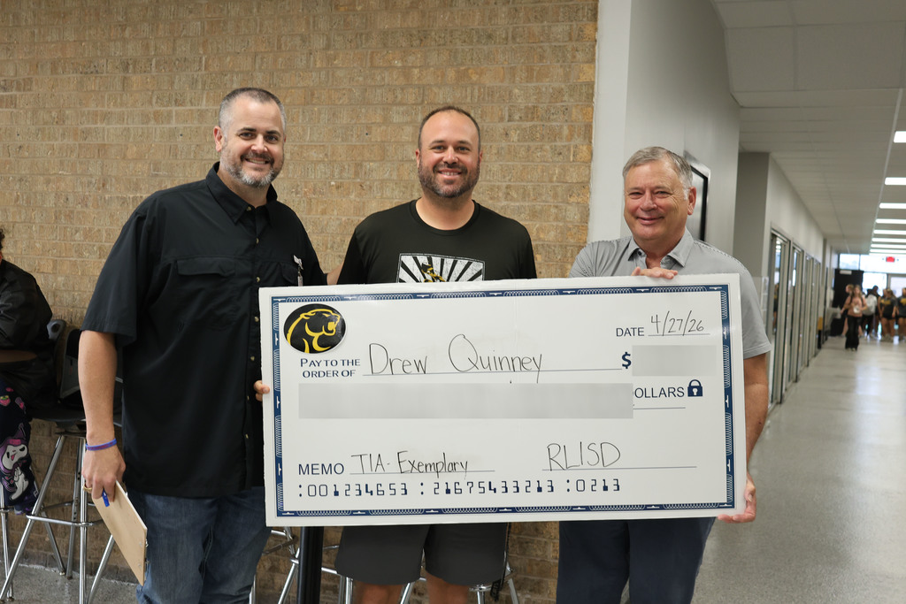 Three people stand in a school hallway holding a large, novelty check. The mood is celebratory and positive, with colorful decorations and student artwork on the walls.