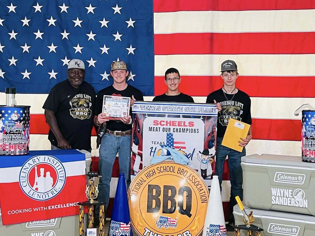 Four people stand proudly in front of an American flag backdrop at a BBQ competition. Trophies and banners surround them. The mood is celebratory.