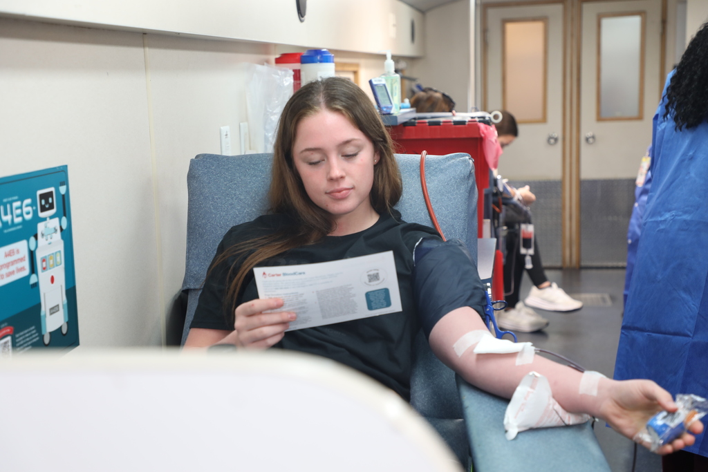 A young woman sits in a hospital chair reading a document while a blood donation process is in progress.