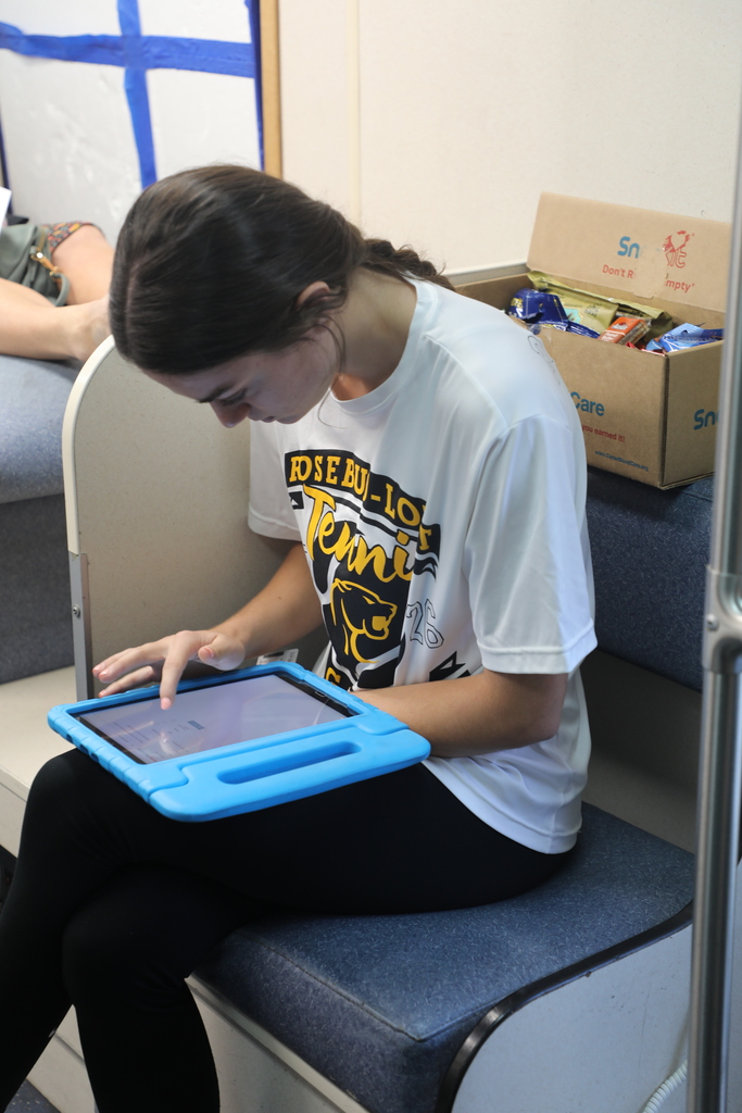 Woman seated on a blue bench, using a tablet with a blue case. Behind her, a cardboard box.