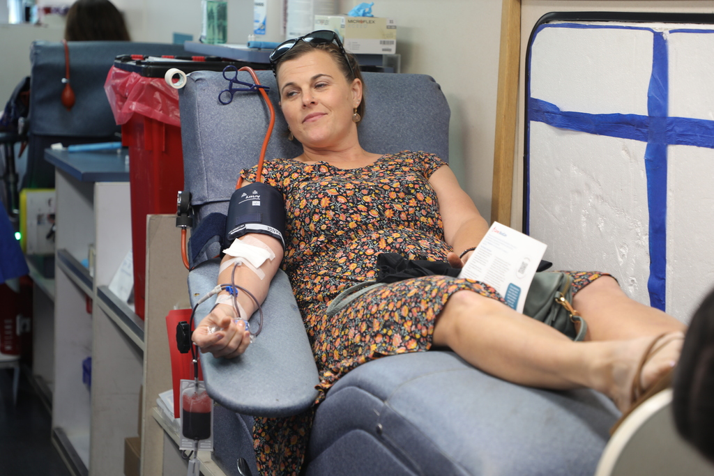 A woman is sitting in a chair with a blood donation setup on her arm.