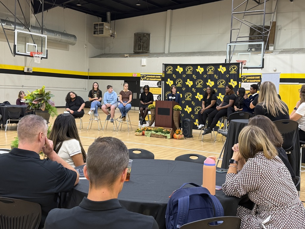 Group of people sitting at tables in a gymnasium. Several are seated at tables; others are standing at a podium.