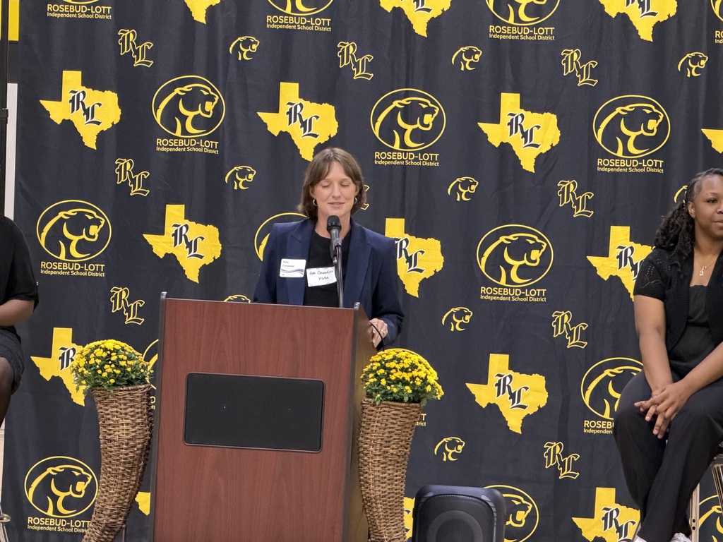 A woman speaks at a podium in front of a black backdrop with yellow designs. Two people sit behind her.