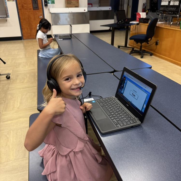 little girl sitting at computer with headphones on giving thumbs up