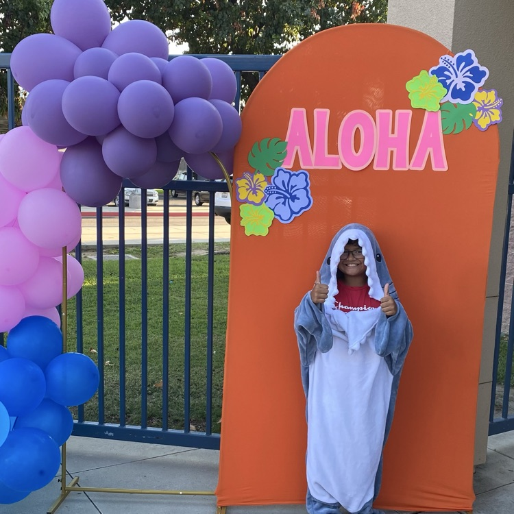 student in a shark costume in front of a sign “Aloha” with purple, pink and blue balloons
