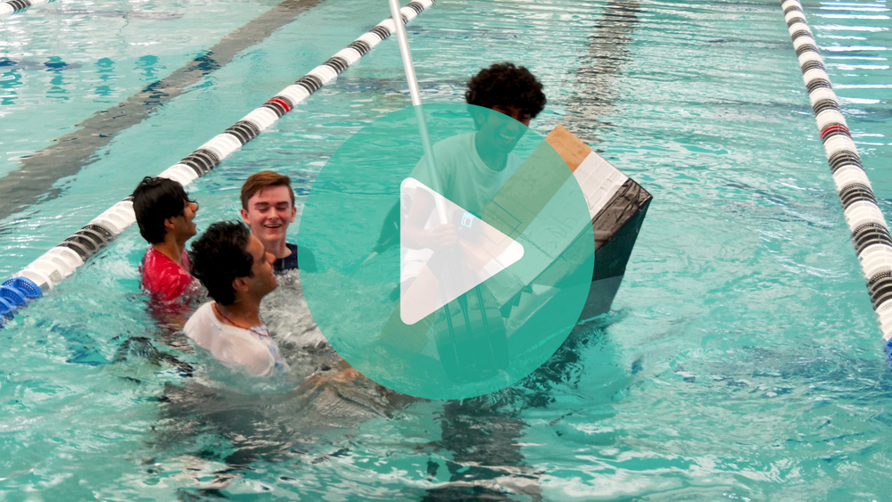 Students in a cardboard boat in the water. The boat is starting to tip over and all the students laugh.