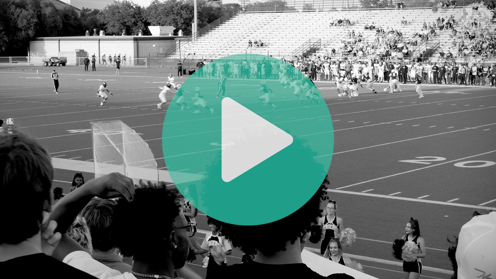 black and white photo sitting in the stands of a high school football game looking onto the field as a football play happens.