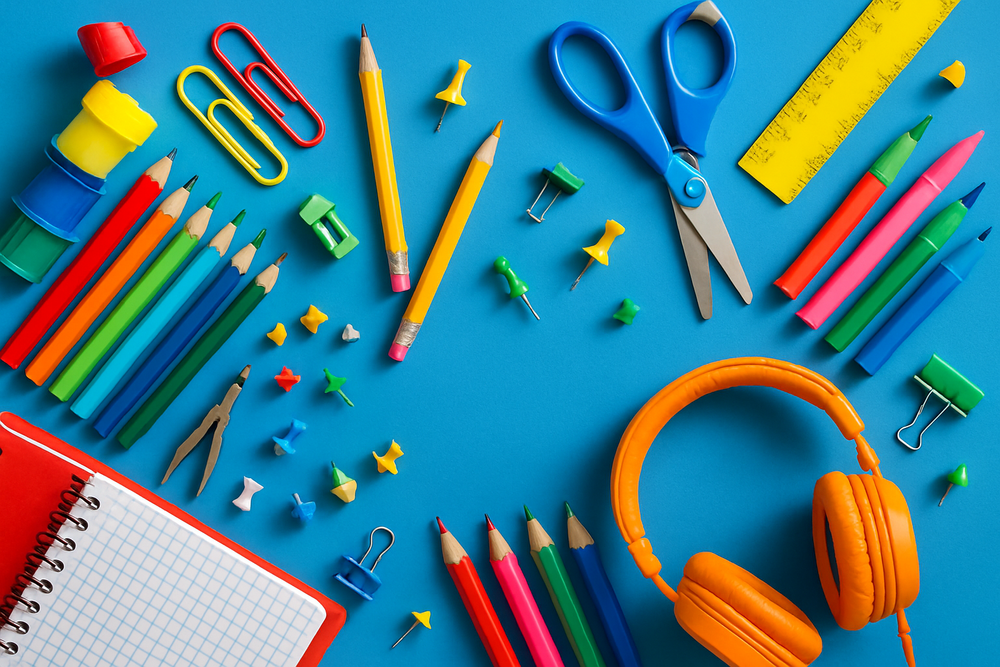 A colorful assortment of school supplies arranged on a bright blue background. Items include sharpened pencils, colored markers, crayons, push pins, a ruler, scissors, a notebook, a pencil sharpener, and a pair of orange headphones, all spread out in an energetic and playful layout.