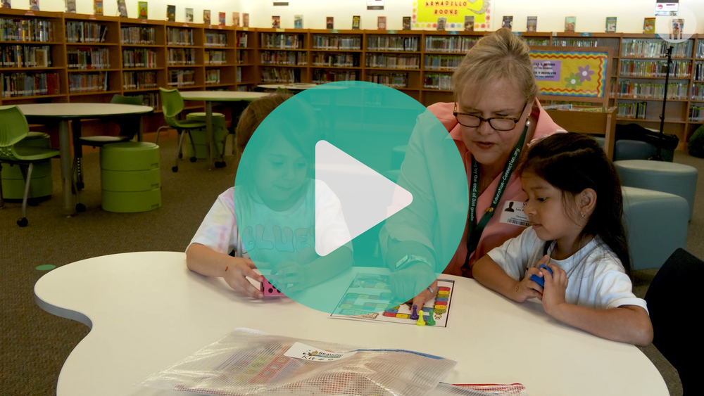 Picture of volunteer helping two students play a word game in the library. 
