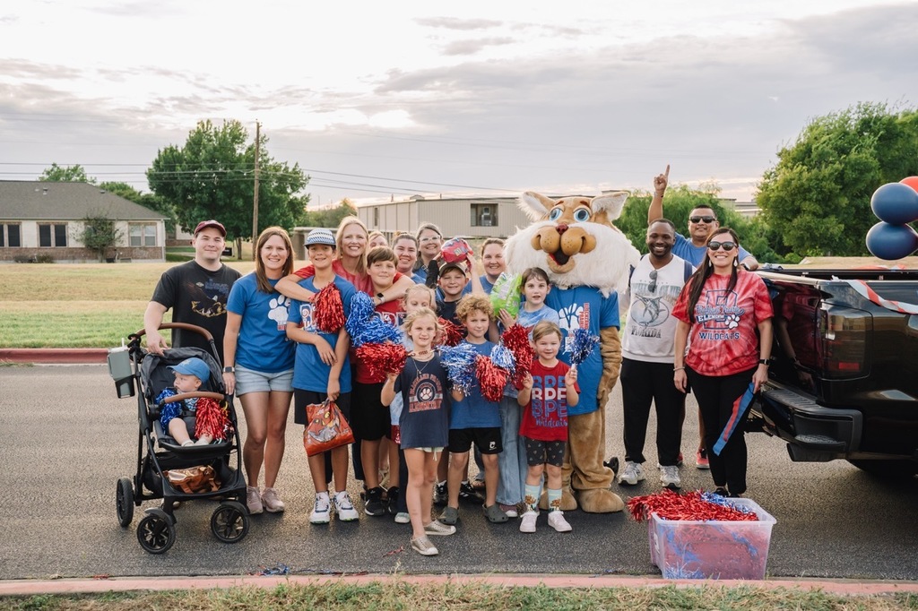 A large group of Blackland Prairie Elementary students, families, and the Wildcat mascot pose for a group photo outdoors before the start of a parade.